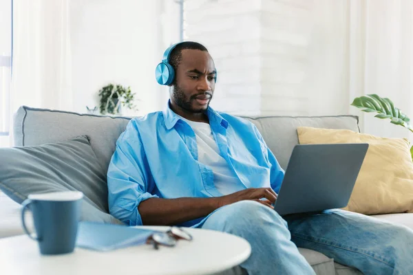 ワイヤレスヘッドフォンを着用し ラップトップを使用している黒人男性は 自宅でソファに座って 自宅からオンラインで働いています アフリカ系アメリカ人の男がコンピューターとサーフィンを使って — ストック写真