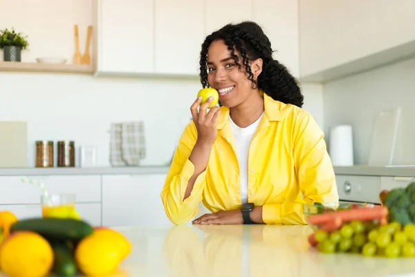 Happy attractive black woman eating fruits, sitting on table in kitchen and smiling at camera, free space. Beautiful lady enjoying eating apple, healthy foods for health care wellness to lose weight