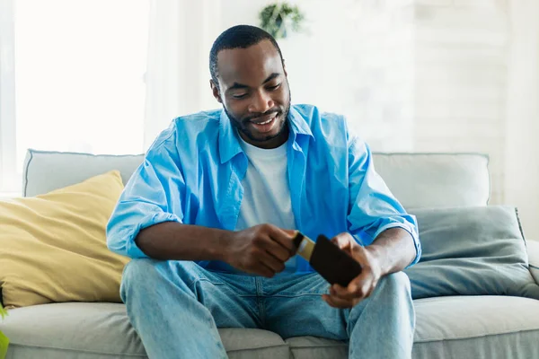 快乐的非洲裔美国人一边笑着从钱包里拿出信用卡 一边坐在客厅的沙发上 免费复印 电子货币概念 — 图库照片