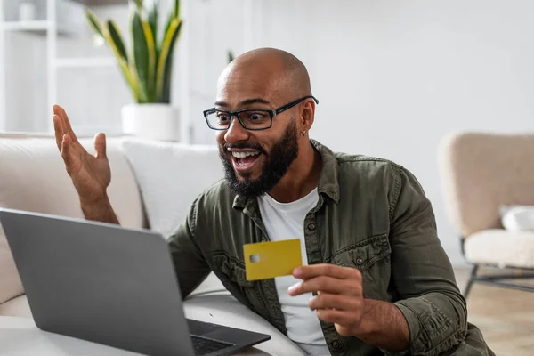 Excited latino man holding credit card in front of laptop, paying for goods on web, shopping on huge sale in web store from home. Cool online promo concept