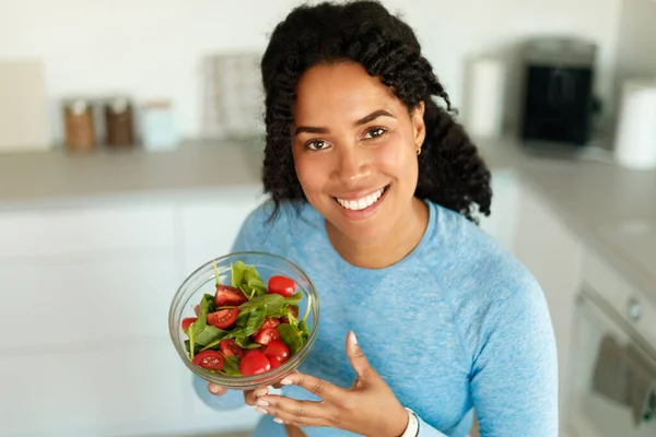 stock image Happy african american woman in sportswear eating healthy salad after domestic workout, sitting in kitchen and smiling at camera. Positive lady holding bowl with fresh vegetables