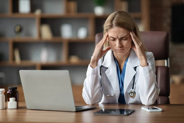 Portrait Of Stressed Female Doctor In Uniform Suffering Headache At Workplace, Sick Overworked Physician Woman Sitting At Desk With Laptop And Touching Temples, Having Acute Migraine, Free Space