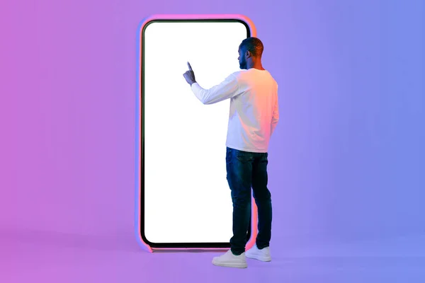 时尚的非洲裔美国人 站在巨大的智能手机旁边 触摸着空白的白屏 使用漂亮的移动应用程序或上网 复制空间 霓虹灯工作室背景 — 图库照片