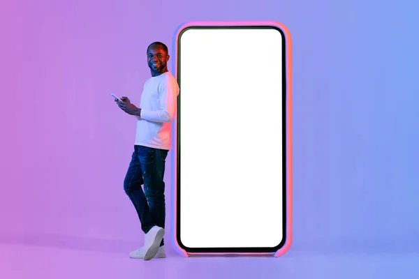 stock image Happy mature african american man in casual outfit standing next to huge cell phone with white empty screen, using smartphone, enjoying newest mobile app, mockup, neon background, copy space