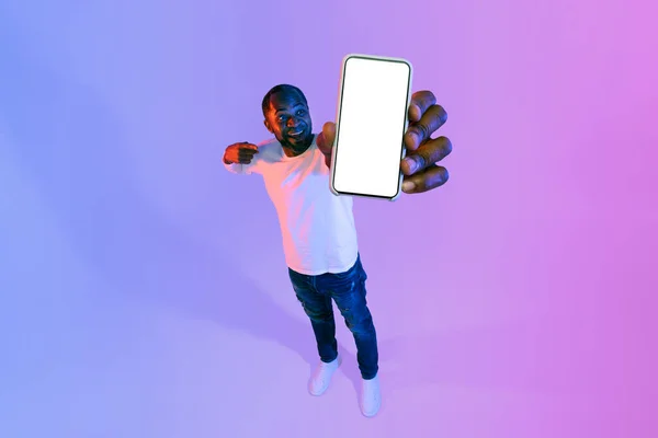 兴奋的中年黑人男子 漫不经心地指着智能手机 手里拿着白色的空白屏风 面带微笑 高角镜头 霓虹灯背景 复制空间 — 图库照片