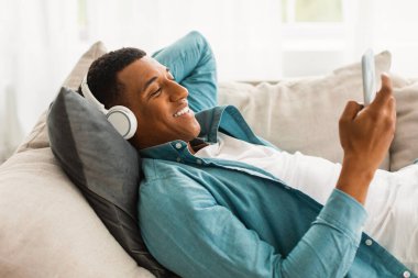 Kablosuz kulaklıklı, akıllı telefonuyla yazı yazan, hafif koltukta uzanan, odada dinlenmekten hoşlanan Afro-Amerikalı genç bir adam. Ses uygulaması, covid-19 salgını sırasında evde favori müzikle rahatlayın