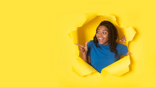 フリースペースで指を指差す興奮アフリカ系アメリカ人女性は 背の高い黄色い紙の背景を通して探してポージング すごい広告コンセプトを提供します パノラマ — ストック写真