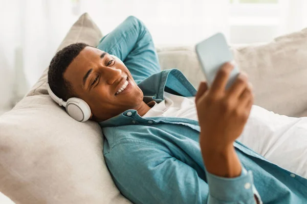 100万人ものアフリカ系アメリカ人の男がワイヤレスヘッドフォンでソファに横になり 電話をかけて リビングルームのインテリアでビデオを見ています 自宅で音楽と休息とリラックス チャットやブログ オーディオアプリ Covid — ストック写真