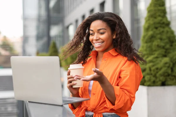 カジュアルな笑顔の若いアフリカ系アメリカ人女性は 街の通りでコーヒーカップをテイクアウトしてノートパソコンでビデオ通話をしています リモートで会う フリーランス ビジネス 技術屋外との仕事と通信 — ストック写真