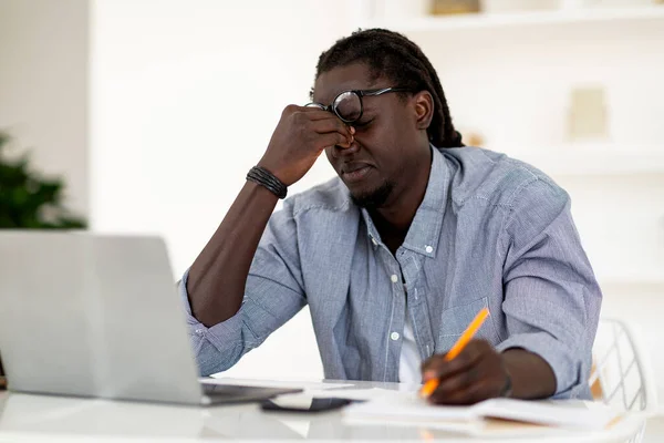 Tired Young Black Man Suffering Eye Strain While Working With Laptop At Desk, Stressed African American Male Massaging Nosebridge And Frowning, Having Migraine Or Eyes Fatigue, Free Space