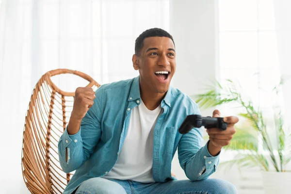 高兴兴奋的千年非洲裔美国人 带着操纵杆玩网络游戏 喜悦胜利 坐在白色客厅的椅子上 游戏者喜欢游戏装置 做出成功的姿态并赢得比赛 — 图库照片