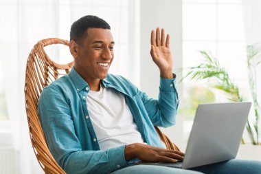 Neşeli bin yıllık Afro-Amerikan erkek el sallıyor, video çağrısı yapıyor, beyaz oturma odasının iç kısmında sandalyede oturuyor. Covid-19 karantinası nedeniyle evdeki iletişim ve toplantı teknolojisi.
