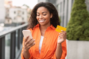 Gülümseyen güzel Afro-Amerikan kadın akıllı telefonuyla yazarak, kredi kartı tutarak, şehir caddesinde satıştan zevk alarak. Finans, banka kumandası, çevrimiçi alışveriş ve iş için para uygulaması