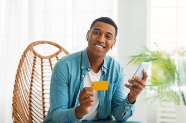Çevrimiçi alışveriş. Milenyum Afrikalı Amerikalı, kablosuz kulaklıklı, akıllı telefon ve kredi kartıyla oturma odasındaki sandalyede oturan adam. Büyük satış, finans, bankacılık ve düzen.