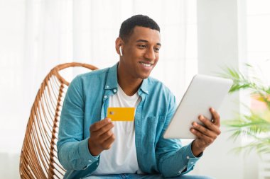 Bin yıllık Afro-Amerikan erkeklerinin kablosuz kulaklıklı, tabletli ve kredi kartlı olmalarına, online alışverişin tadını çıkarmalarına, beyaz oturma odasının içindeki sandalyeye oturmalarına sevindim. Alışveriş ve satış, reklam ve teklif