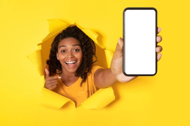 Elinde Smartphone olan neşeli Afro-Amerikan kadın başvuru ilanı için boş ekranı göstererek yırtık kağıtta delik pozu veriyor. Harika bir mobil teklif. Model
