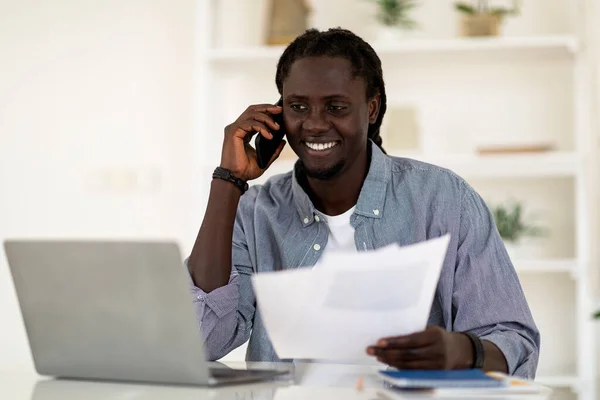 Smiling Black Man Talking On Cellphone And Working With Documents While Sitting At Desk At Home Office, Young African American Male Freelancer Using Laptop Computer And Checking Papers, Copy Space
