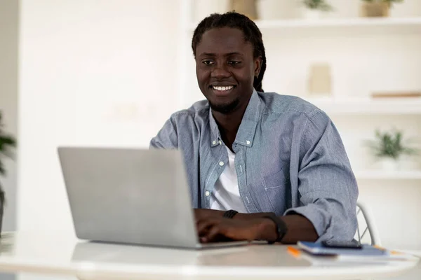 Freelance Career. Portrait Of Young Smiling Black Man Working With Laptop While Sitting At Desk In Home Office, Happy Millennial African American Man Using Computer For Remote Job, Free Space