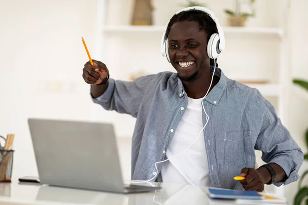 自宅でラップトップで音楽を聞いている間に楽しい時間を過ごしている陽気な黒人男性 コンピュータで机に座って仮想ドラムを演奏する幸せな若いアフリカ系アメリカ人男性 フリースペース — ストック写真