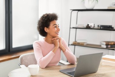 Kıvırcık saçlı, gülümseyen Afro-Amerikan kadın ofis içi dizüstü bilgisayarı olan boş bir masaya bakar ve düşünür. İş, serbest çalışma, uzaktan çalışma, sosyal uzaklık ve yeni normal
