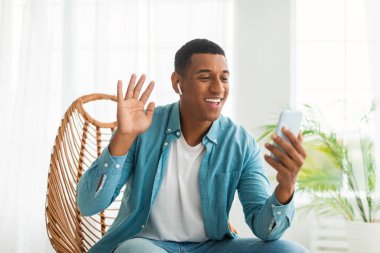 Bin yıllık neşeli Afro-Amerikan erkek, kablosuz kulaklıklı, el sallayan, video çağrısı yapan, beyaz oturma odasının iç tarafında oturan. Uzaktan buluşma, sohbet ve iletişim uygulaması