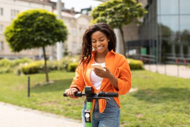 Elektrikli scooterlı, akıllı telefondan yazı yazan mutlu, Afro-Amerikalı kadın yazın şehirde eğleniyor. Harika hizmet, yeni uygulama, tek başına trafik ve ekoloji taşımacılığı.