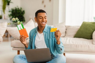 Gelecekteki yolculuğun siparişi. Kredi kartı, pasaport ve uçak biletleri olan mutlu, Afro-Amerikalı bir adam oda içinde bilgisayar kullanıyor. Cashback, seyahat, tatil ve covid-19 salgınından sonra seyahat