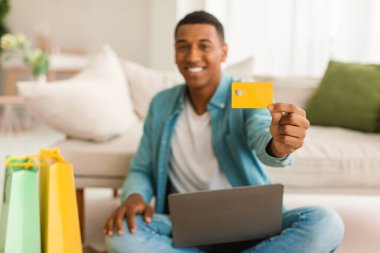 Kredi kartı, bilgisayar ve alışveriş torbaları olan neşeli, Afro-Amerikalı genç bir adam oturma odasının içinde online alışveriş yapmaktan hoşlanıyor, bulanık. Nakit para, alış veriş manyağı için ev yapımı covid-19.