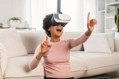 VR gözlüklü kıvırcık saçlı mutlu Afro-Amerikalı bayan oturma odasının iç tarafındaki boşluğa elleriyle dokunuyor. Modern teknoloji ve sanal gerçeklikle evde eğlence