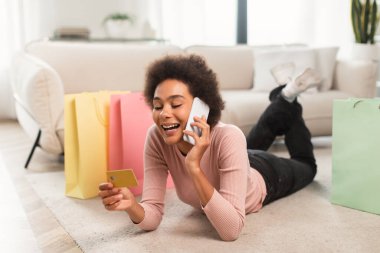 Kıvırcık saçlı mutlu Afro-Amerikan bayan yerde yatıyor, akıllı telefonuyla açık oturma odasında online alışveriş yapıyor. Bankacılık ve para, uzaktan satın alma emri