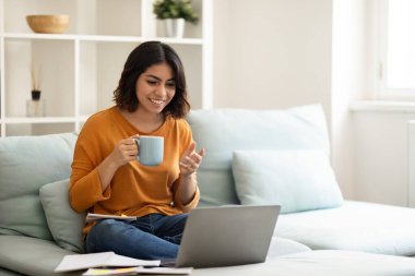 Çevrimiçi İletişim. Mutlu Arap kadın laptopta video görüşmesi yapıyor ve evde kahve içiyor, arkadaşlarıyla telekonferans yapan ve sıcak içeceğin tadını çıkaran neşeli Orta Doğulu kadın, boşluğu kopyalıyor.