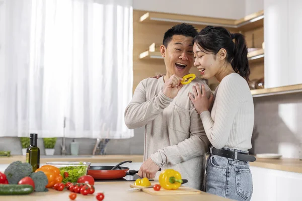 stock image Handsome japanese middle aged man cooking dinner for his beautiful young wife at home, asian chef feeding his slender girlfriend bell pepper while preparing delicious healthy meal, copy space