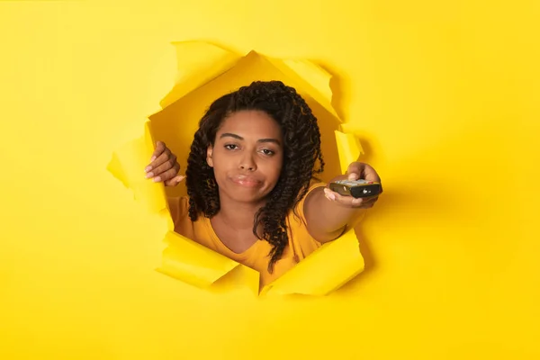 不满的非洲裔美国妇女看电视开关渠道指点遥控装置通过孔在扭曲的黄色纸片 工作室拍摄 无聊的电影概念 大学生活 — 图库照片