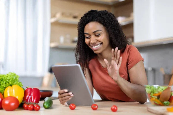 Happy young black lady in a red t-shirt waving at a tablet has a video call at a table with limited vegetables in a modern kitchen interior. meeting new app, food blog, work from home, new normal and