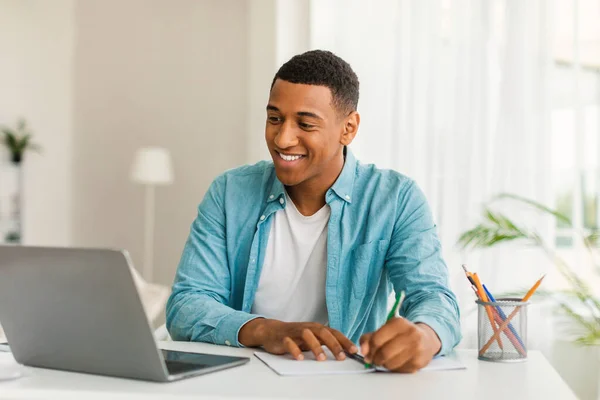 部屋やオフィスのインテリアでノートを作るためのカジュアルな使用コンピュータで幸せなハンサムな若いアフリカ系アメリカ人の男性マネージャー ビジネスとフリーランスリモートで 家庭での社会的距離Covid — ストック写真