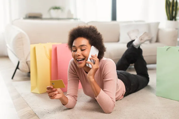 幸せな若いアフリカ系アメリカ人女性は 巻き毛の床に位置し クレジットカードを使用し 光のリビングルームのインテリアでオンラインショッピングのためのスマートフォンで呼び出します 銀行とお金 遠隔注文を購入する — ストック写真