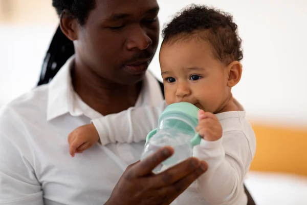 Caring African American Father Giving Water Bottle To His Cute Little Baby, Thirsty Adorable Black Infant Boy Drinking Water From Sippy Cup With Straw While Relaxing In Dads Arms, Closeup Shot
