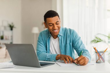 Happy busy millennial african american man with laptop works and makes notes at table in living room interior. Tutoring, education, study, freelance and new normal at home during covid-19 quarantine clipart