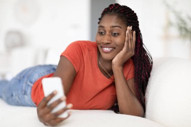 Gülümseyen siyah kadın evdeki rahat koltukta akıllı telefonuyla rahatlıyor, mutlu Afrikalı genç Amerikalı bayan modern Gadget ile koltukta dinleniyor, cep telefonuyla interneti tarıyor,