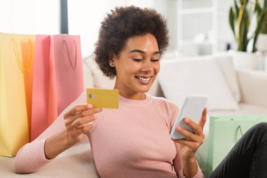 Kıvırcık saçlı, çantalı neşeli Afro-Amerikan bayan, açık oturma odasının içinde online alışveriş yapmak için kredi kartı ve akıllı telefon kullanıyor. Finans, bankacılık ve para uygulamalı sipariş, büyük satış.