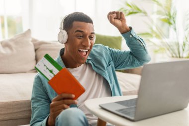 Kablosuz kulaklıklı, elinde pasaport ve uçak biletleri olan, bilgisayara bakarak zafer ve başarı jesti yapan genç, heyecanlı siyah bir erkek. Tatilden, yolculuktan ve covid-19 'dan sonra seyahatten duygular