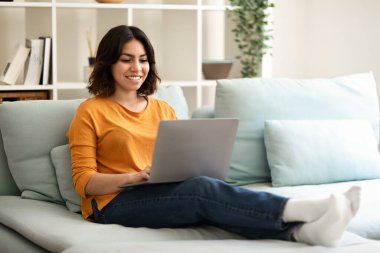 Gülümseyen Orta Doğulu genç bir kadın dizüstü bilgisayarında internete göz gezdirirken evinde dinlenirken mutlu bir Arap kadını rahat bir oturma odasında oturuyor ve bilgisayarda boşluğu kopyalıyor.