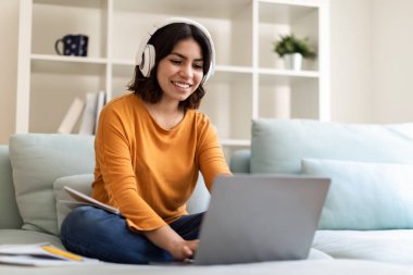 Mesafe öğrenme. Kulaklıklı mutlu bir genç Arap kadın evde dizüstü bilgisayarla ders çalışıyor, orta doğu kadınlarının oturma odasında koltukta otururken online eğitim için bilgisayar kullanmalarına gülüyor.
