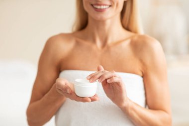 Elinde nemlendirici krem bulunan kavanozu tutan tanınmayan olgun bir kadın, beyaz havlu giymiş bir kadın ve evde güzel cilt bakımı yapıyor.