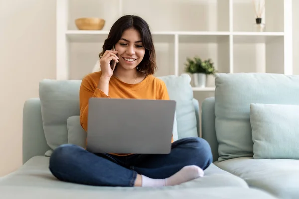 漂亮的中东年轻女性 使用笔记本电脑 在家里谈论手机 微笑着坐在沙发上的阿拉伯女性 在电脑上浏览互联网 享受愉快的电话交谈 — 图库照片