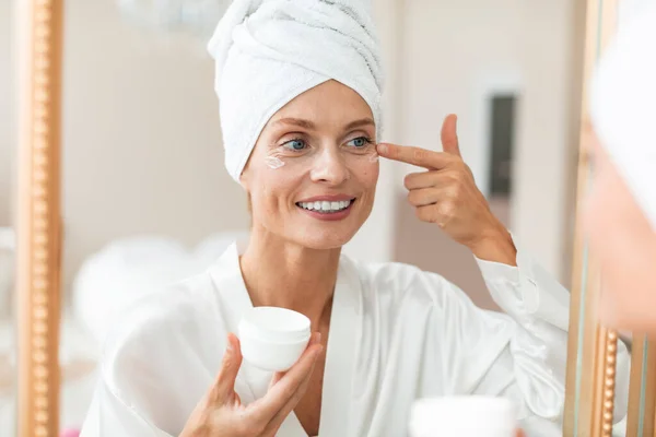 美しい中年女性で絹のようなバスローブに保湿アイクリームを適用し 鏡を見て 顔のケアルーチンを楽しんで バスルームインテリア — ストック写真