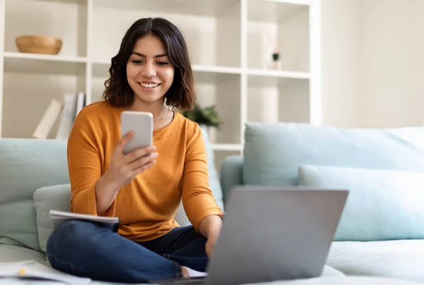 用智能手机和笔记本电脑在家中沙发上放松的年轻中东女性 在手机上浏览新的应用 在电脑上浏览互联网 复制空间 — 图库照片