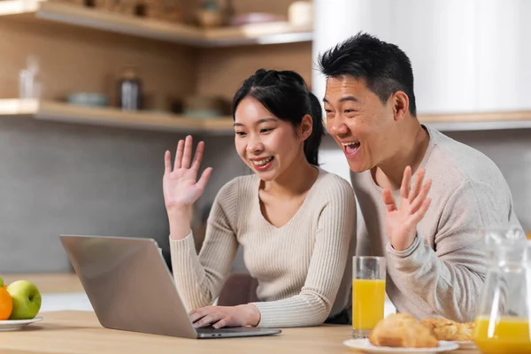 stock image Happy chinese couple pretty young woman and middle aged man sitting at kitchen desk, having video conference while enjoying healthy breakfast, looking at laptop screen and waving, copy space