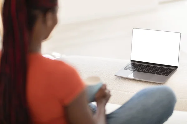 Black Woman Using Blank Laptop With White Screen And Drinking Coffee At Home, Over Shoulder View Of Young African American Female Relaxing With Computer In Living Room, Mockup Image With Copy Space