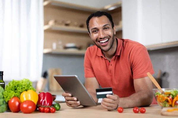 Smiling young black male in red t-shirt with tablet and credit card at table with organic vegetables at kitchen interior. App for online shopping, new normal, ordering goods, finance and banking